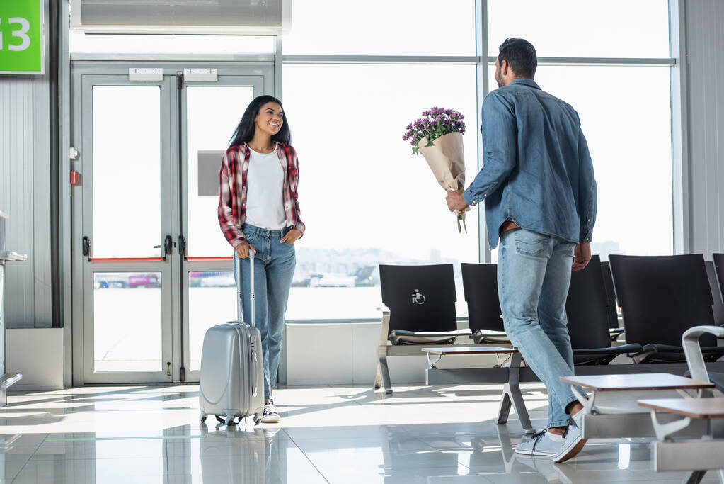 Neşeli Afrikalı Amerikalı kız arkadaşıyla havaalanında buluşurken elinde çiçek tutan bir adam. - Fotoğraf, Görsel
