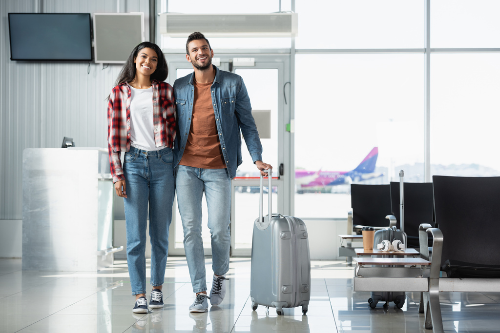 heureux couple multiculturel avec bagage marche dans l'aéroport moderne  - Photo, image