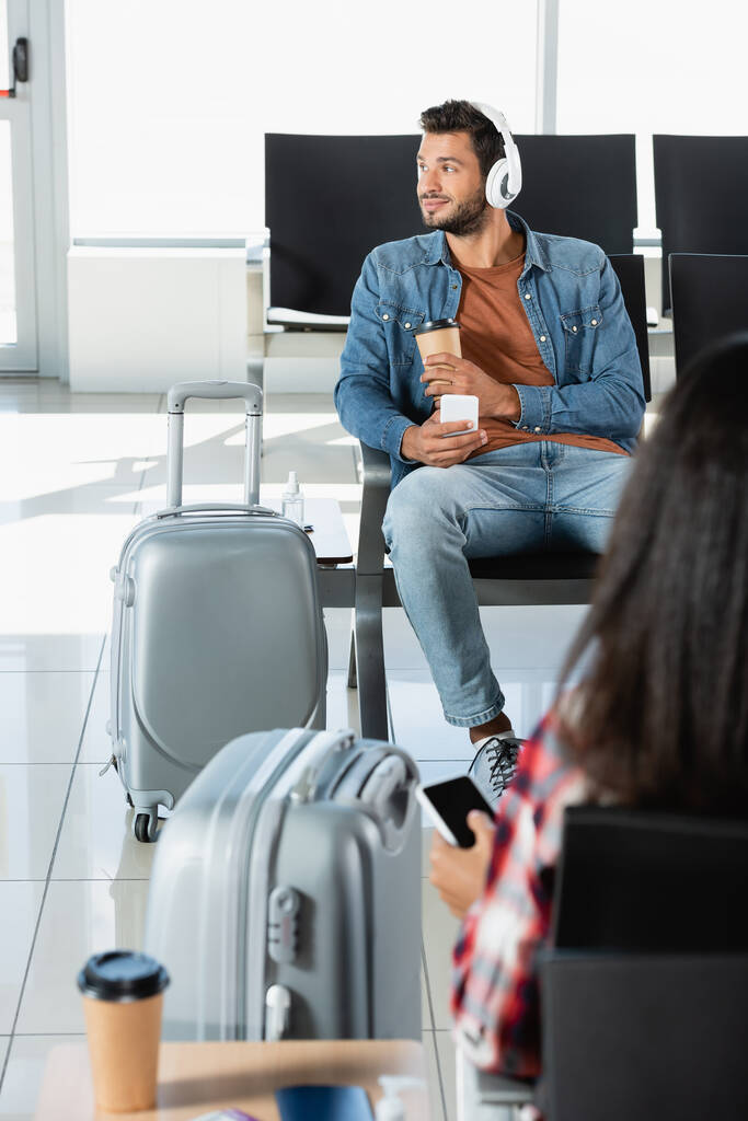uśmiechnięty mężczyzna w słuchawkach trzymający papierowy kubek i smartfon w pobliżu bagażu i kobieta na niewyraźnym pierwszym planie  - Zdjęcie, obraz