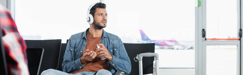 barbudo hombre en auriculares usando desinfectante en el aeropuerto cerca de pasajero en primer plano borrosa, bandera - Foto, imagen