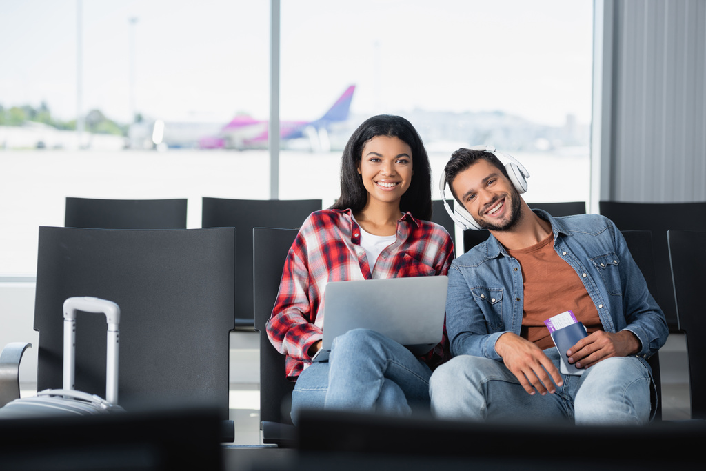 homem feliz ouvindo música perto de mulher americana africana com laptop no aeroporto  - Foto, Imagem
