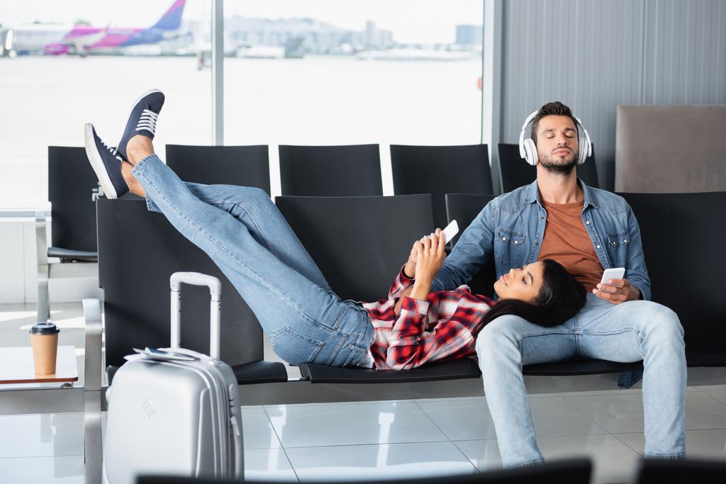 african american woman using phone and lying on bearded man in headphones in departure lounge  - Photo, Image