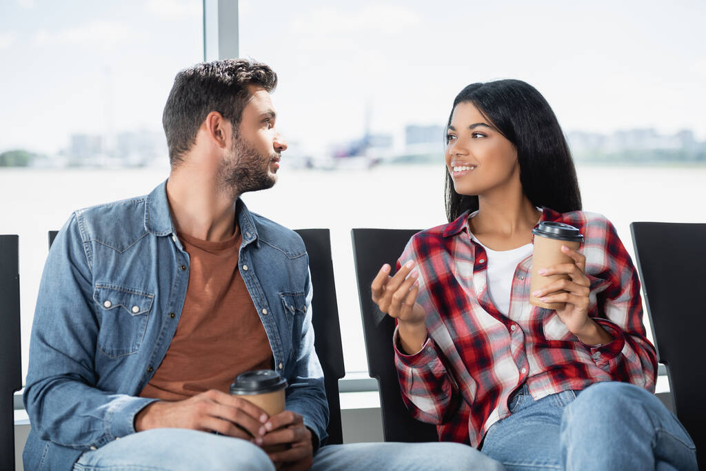 χαρούμενο πολυπολιτισμικό ζευγάρι κρατώντας καφέ για να πάει, ενώ κάθεται στο σαλόνι αναχώρησης του αεροδρομίου  - Φωτογραφία, εικόνα