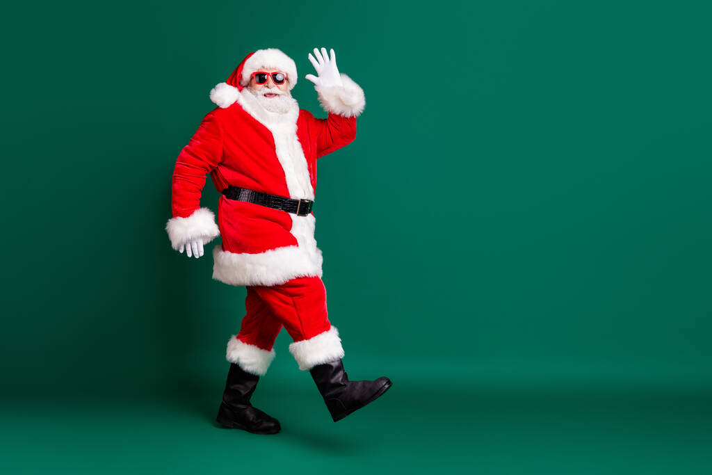 Full body size profile photo of pensioner grandpa walking mall corridors greeting shop customers wear red santa costume coat gloves sunglass headwear isolated green color background - Foto, Imagem