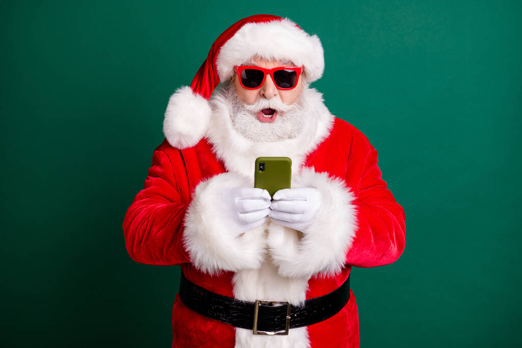 Fénykép a nyugdíjas stílusos nagypapa fehér szakáll tartsa telefon nyitott száj sokkolt váratlan eladás kopás piros mikulás x-mas jelmez kabát kesztyű napszemüveg fejfedő elszigetelt zöld szín háttér - Fotó, kép