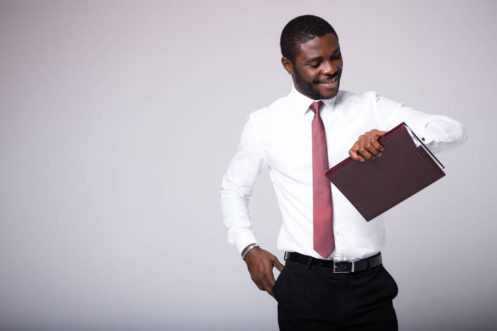 Ένας Αφροαμερικάνος μεσίτης στέκεται με ένα φάκελο στα χέρια του, κοιτάζει το ρολόι του και περιμένει τον πελάτη του. - Φωτογραφία, εικόνα