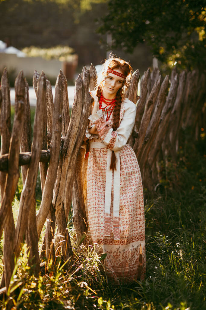 Una hermosa chica eslava con el pelo largo y rubio y los ojos marrones en un traje bordado blanco y rojo se encuentra junto a una cerca de madera.Ropa tradicional de la región de Ucrania.Día de verano - Foto, imagen