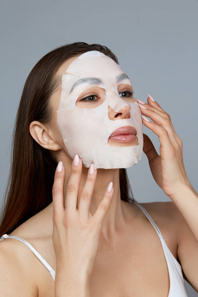 Cuidado de la piel de la cara belleza. Mujer aplicar una máscara hidratante de tela en la cara. Modelo de niña con máscara cosmética. Tratamiento facial - Foto, imagen