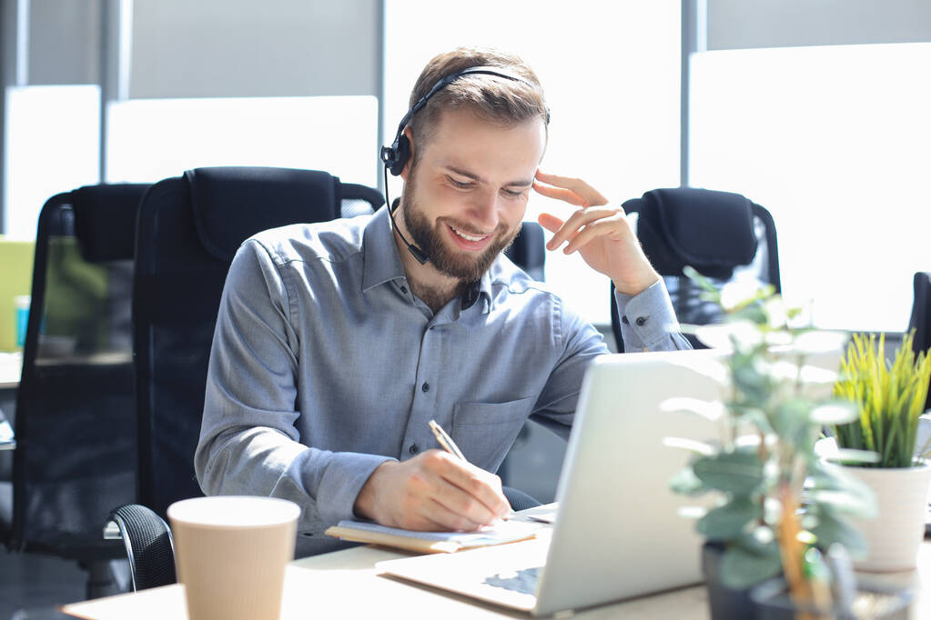 Lächelnder männlicher Call-Center-Betreiber mit Kopfhörern sitzt im modernen Büro, konsultiert Online-Informationen in einem Laptop, sucht Informationen in einer Akte, um dem Kunden zu helfen - Foto, Bild