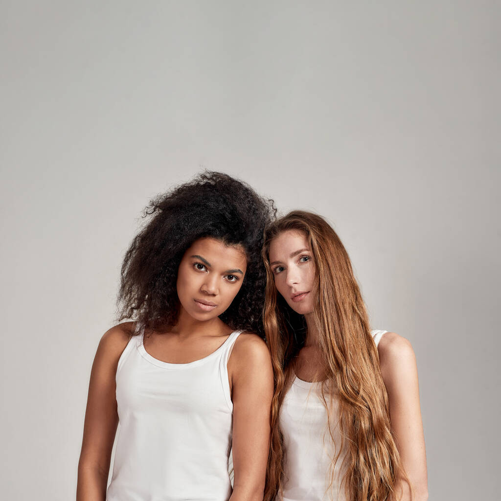 Портрет двох молодих різноманітних жінок у білих сорочках, які дивляться на камеру, стоячи разом ізольовано на сірому фоні
 - Фото, зображення