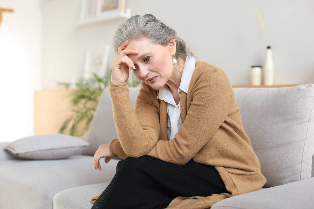 Αγχωμένη μεσήλικη γυναίκα κάθεται στον καναπέ στο σαλόνι, χαμένη στη σκέψη - Φωτογραφία, εικόνα