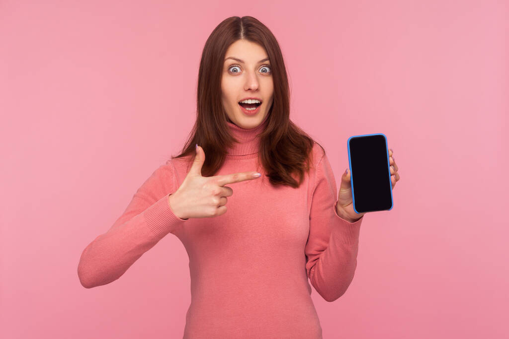 Donna bruna estremamente sorpresa che punta il dito contro lo smartphone in mano guardando la fotocamera con un'espressione stupita e stupita. Indoor studio girato isolato su sfondo rosa - Foto, immagini