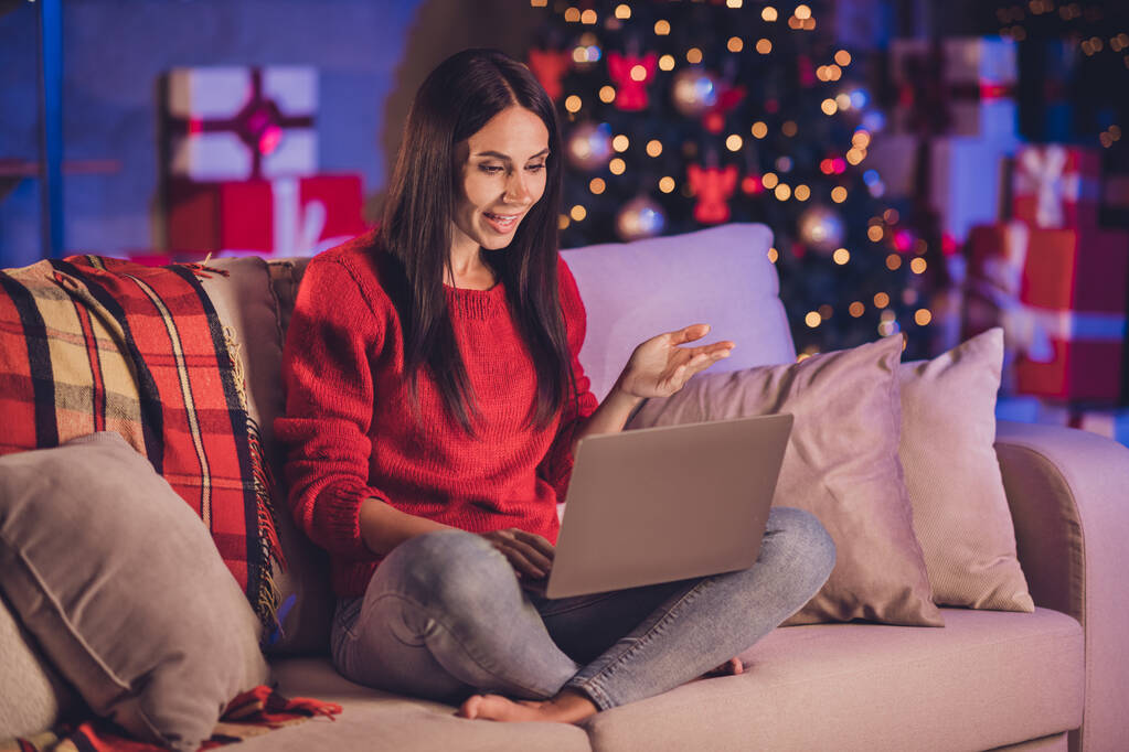 Zdjęcie portret kobiety siedzącej na kanapie w pozycji lotosu z laptopem w czerwonym swetrze rozmawiającej z kamerką internetową z dekoracjami świątecznymi w pomieszczeniach - Zdjęcie, obraz
