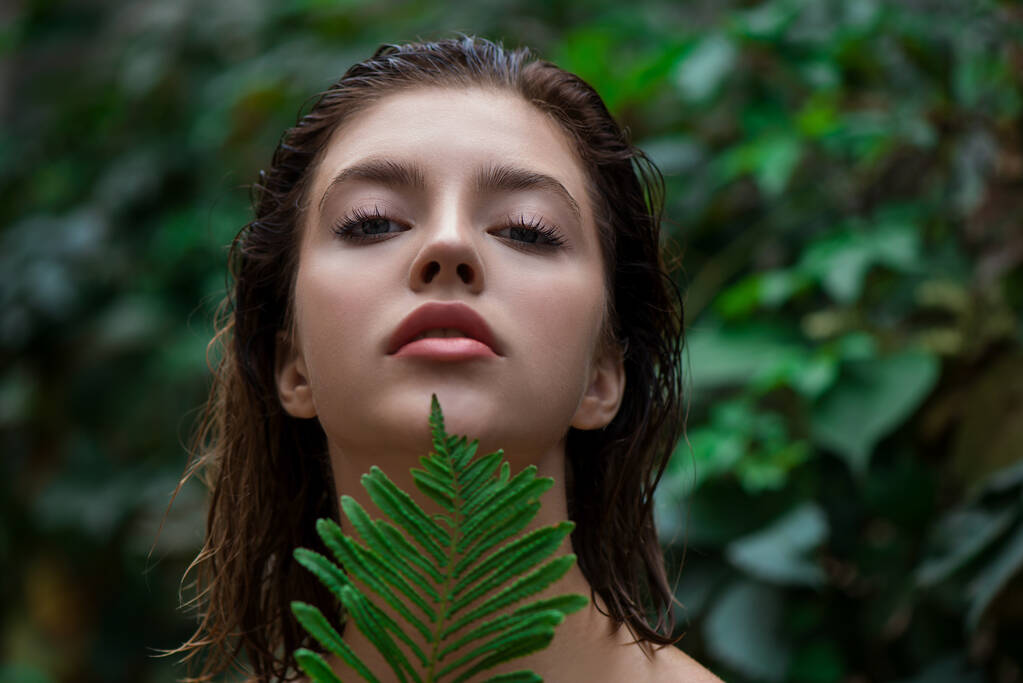 Beautiful young woman with perfect skin and natural make up posing front of plant tropical green leaves background with fern. Teen model with wet hair care of her face and body. SPA, wellness, bodycare and skincare.  Close up, selective focus. - Photo, Image