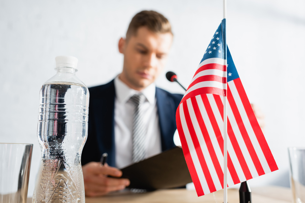 Bottiglia d'acqua e piccola bandiera americana sul tavolo con politico offuscato sullo sfondo - Foto, immagini