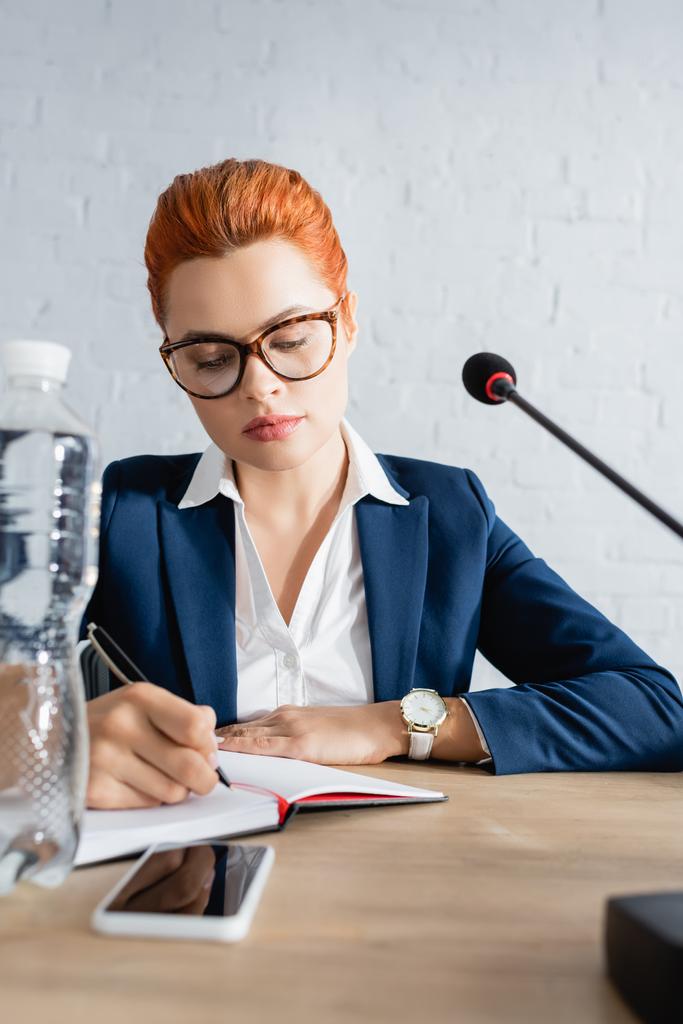 Resmi giyinmiş kızıl saçlı kadın, elinde su ve mikrofonla masada otururken deftere yazı yazıyor. - Fotoğraf, Görsel