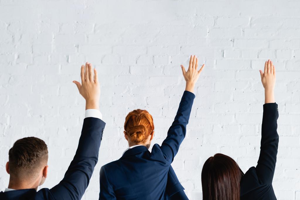 Rückansicht von Frauen und Männern, die mit den Händen in der Luft gegen eine weiße Backsteinwand stimmen - Foto, Bild
