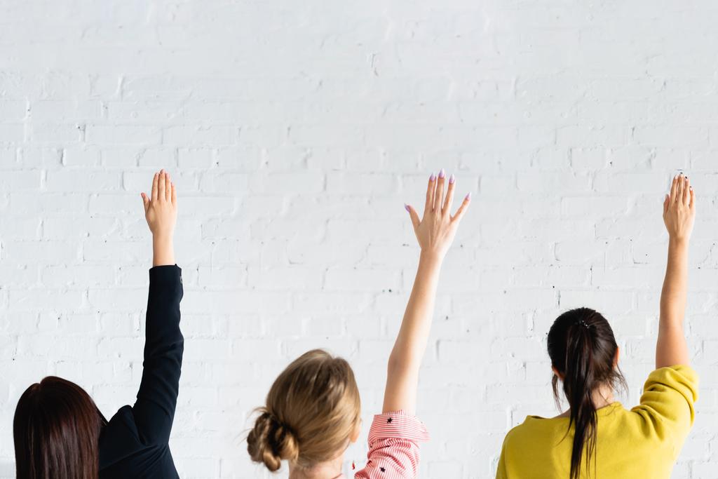 zadní pohled žen hlasujících se zdviženýma rukama proti zdi z bílých cihel - Fotografie, Obrázek