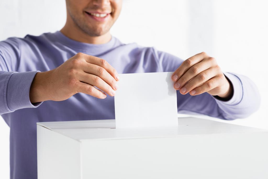 vista recortada del hombre sonriente insertando papeleta en la urna electoral sobre fondo borroso - Foto, imagen