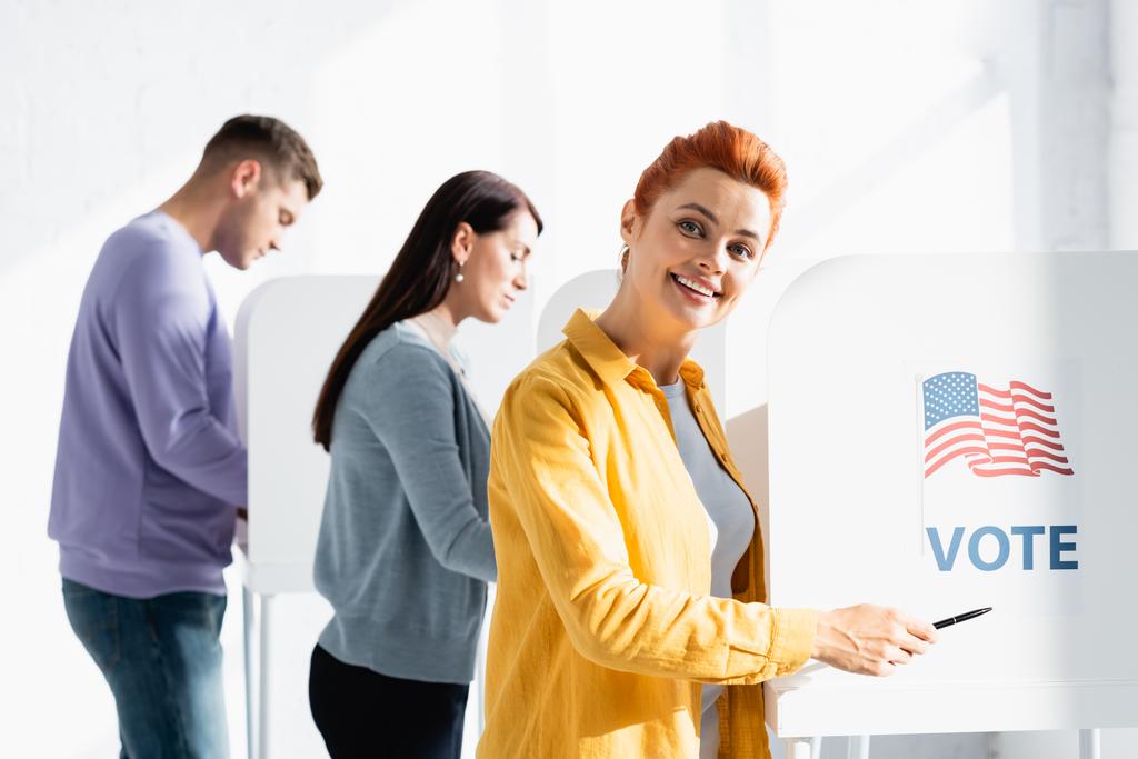 Amerikan bayrağında kalemle işaret eden gülümseyen kadın ve bulanık arka planda oy kullanma kabininde oylama. - Fotoğraf, Görsel