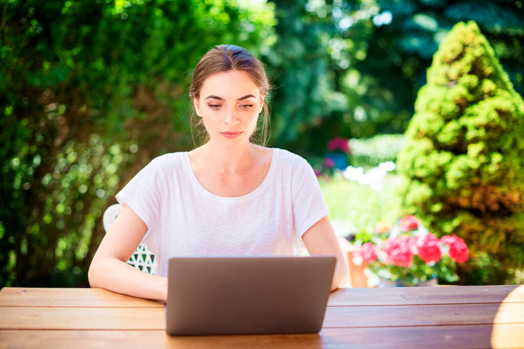 Bella giovane donna che utilizza il computer portatile mentre si siede in giardino e lavora da casa.  - Foto, immagini