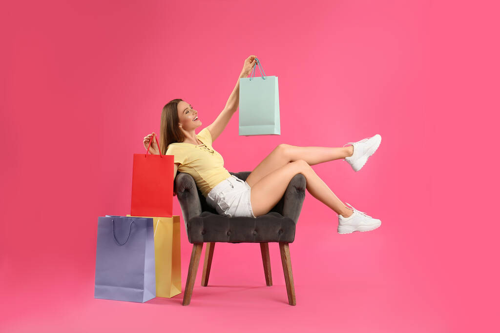 Belle jeune femme avec des sacs à provisions en papier dans le fauteuil sur fond rose - Photo, image