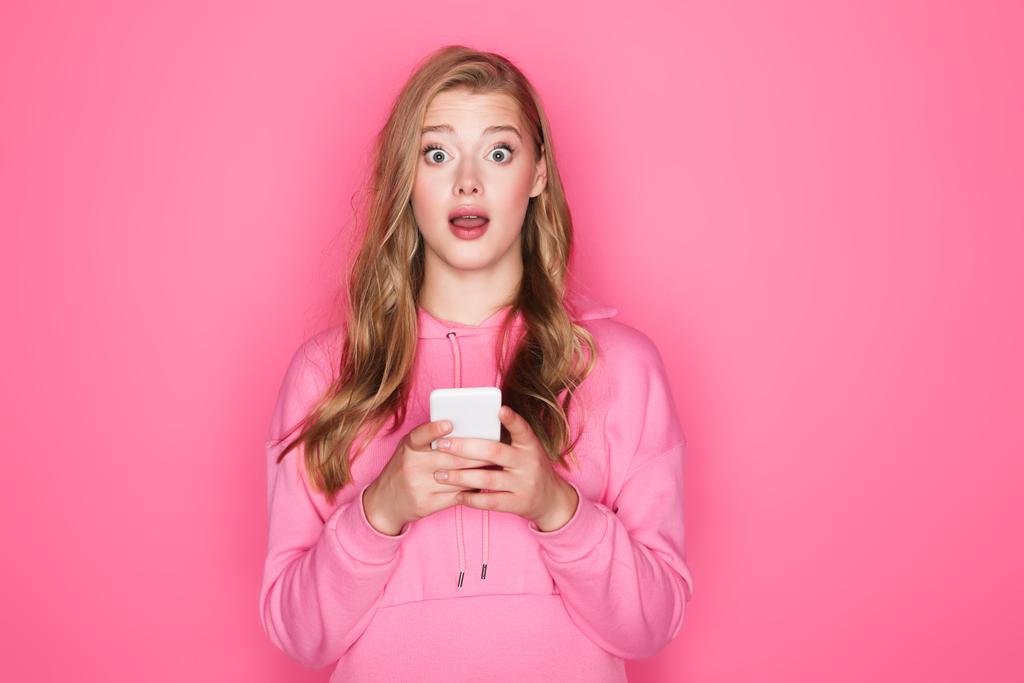 geschokt mooie vrouw met open mond houden smartphone op roze achtergrond - Foto, afbeelding