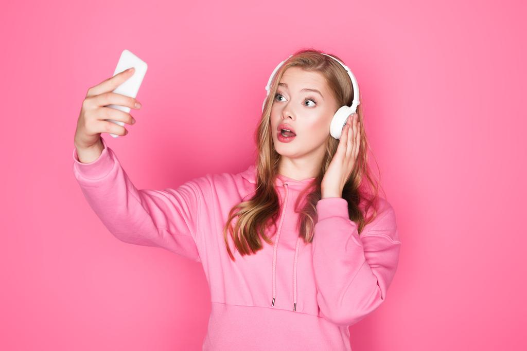 shocked beautiful woman in headphones taking selfie on pink background - Photo, Image