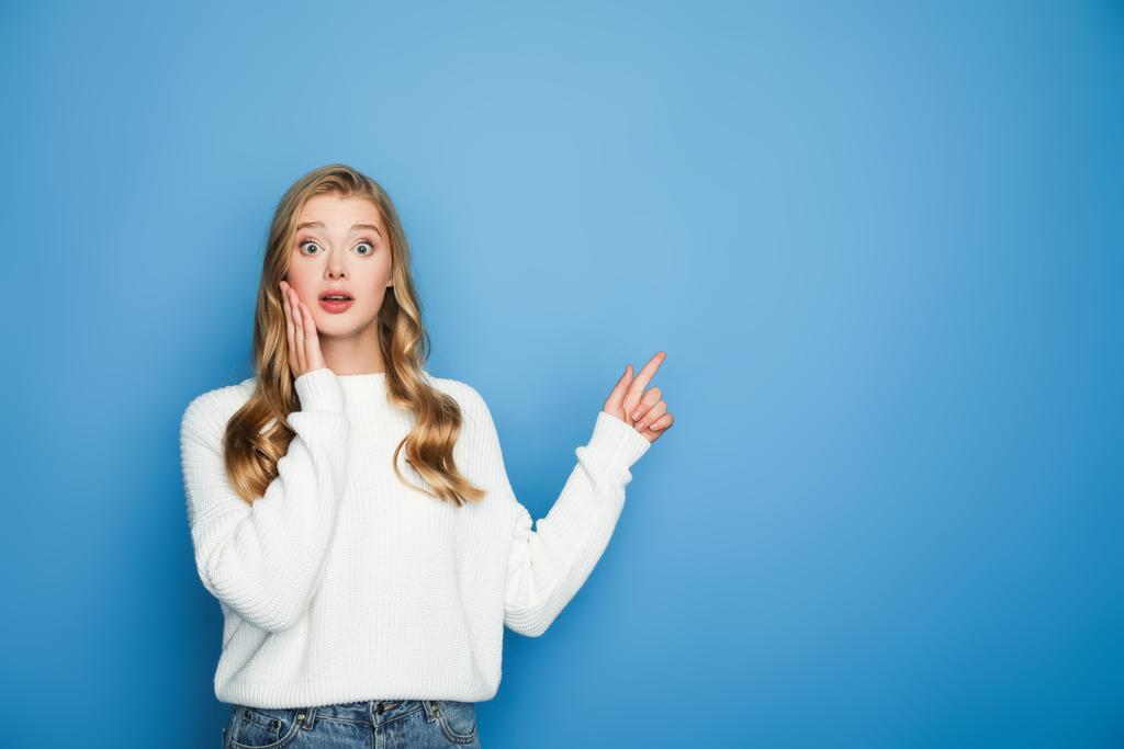 shocked blonde beautiful woman in sweater pointing with finger isolated on blue - Photo, Image
