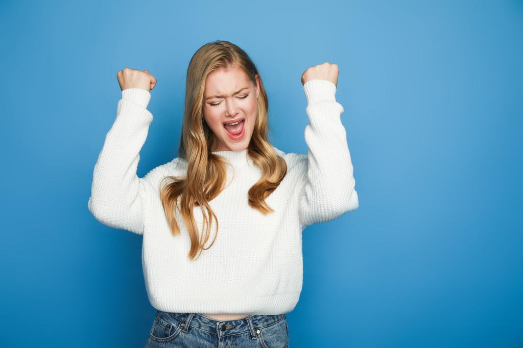 happy blonde beautiful woman in sweater showing yeah gesture isolated on blue background - Photo, Image