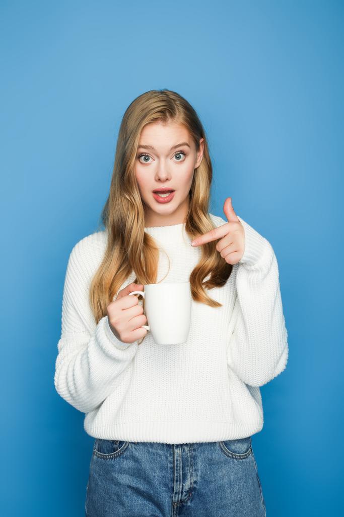 surprised blonde beautiful woman in sweater pointing at mug isolated on blue background - Photo, Image