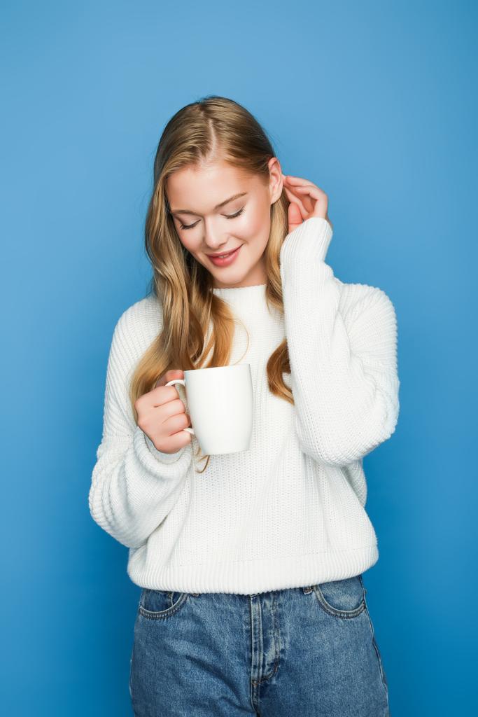 happy blonde beautiful woman in sweater pointing at mug isolated on blue background - Photo, Image