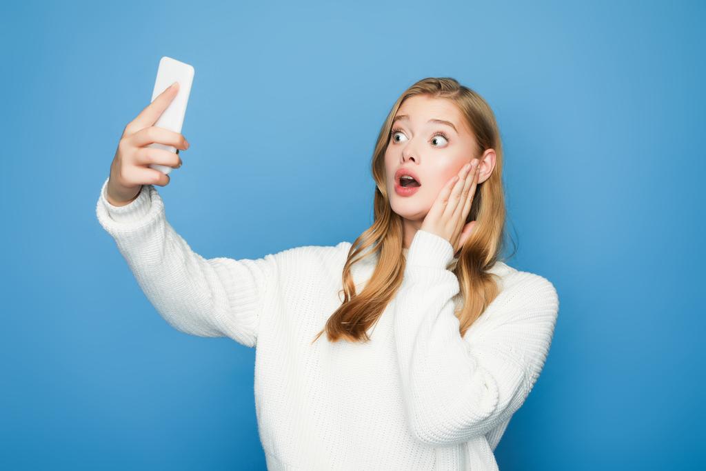 geschokt blond mooi vrouw het nemen van selfie geïsoleerd op blauwe achtergrond - Foto, afbeelding
