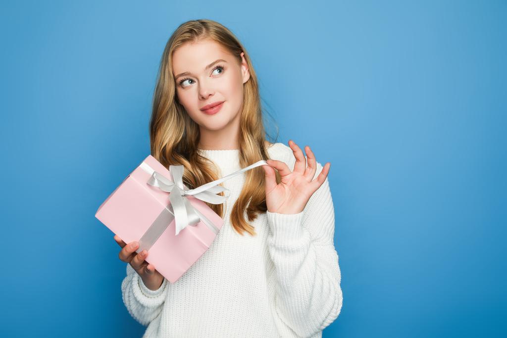 dreamy blonde beautiful woman in sweater with gift box isolated on blue - Photo, Image