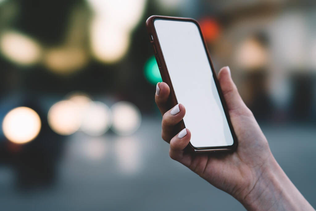 Crop hembra anónima utilizando un teléfono móvil inalámbrico moderno con pantalla táctil en blanco mientras sostiene el dispositivo en la mano sobre fondo borroso de la calle - Foto, imagen