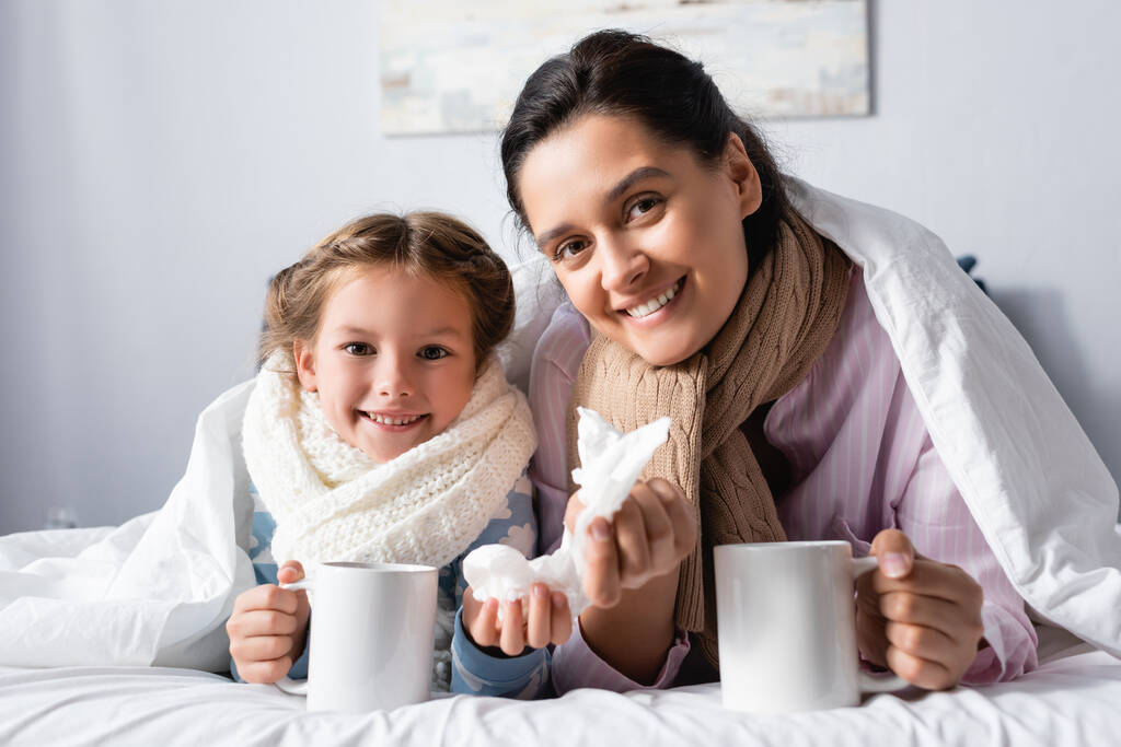 diseased, smiling woman with child holding cups of warm drink in bed - Photo, Image