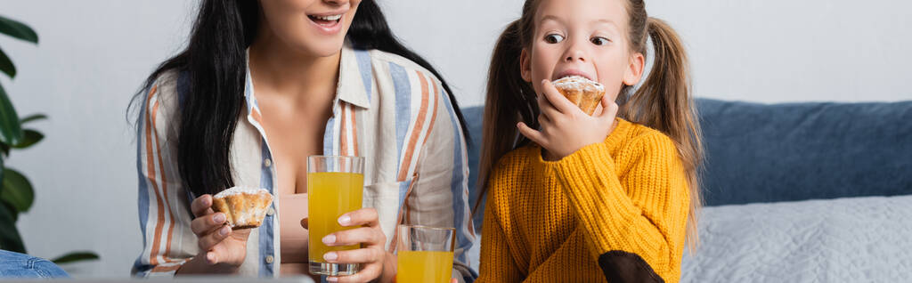 девушка ест вкусный кекс возле улыбающейся матери с апельсиновым соком, баннер - Фото, изображение