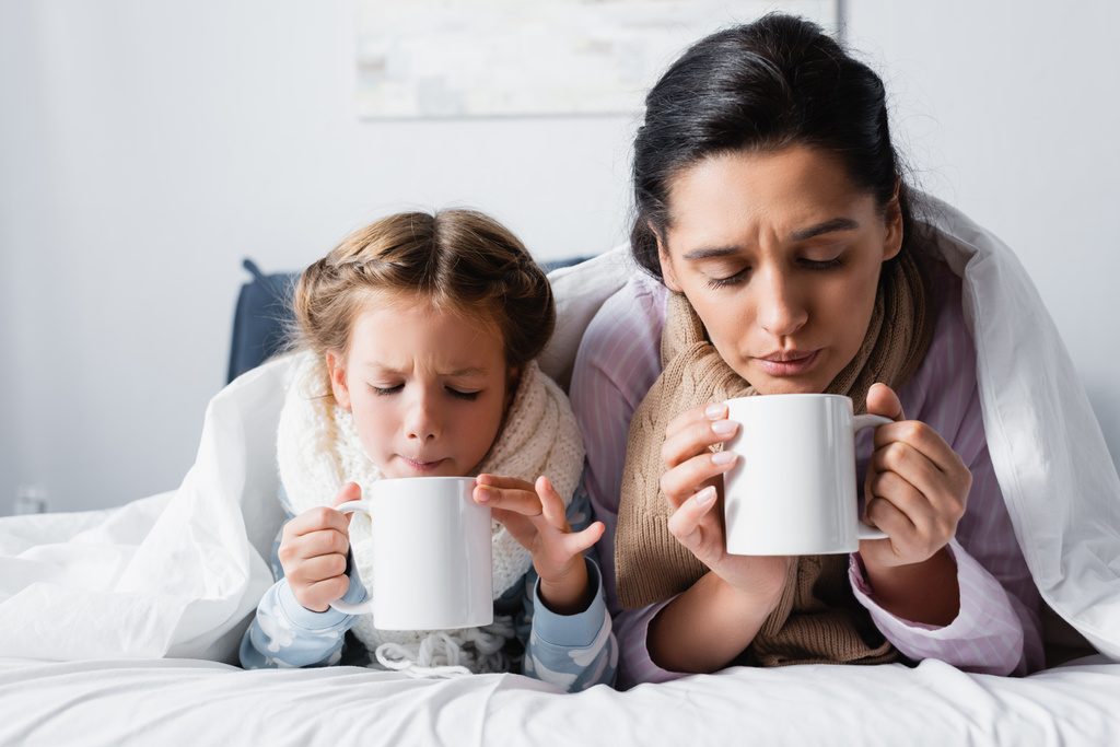 diseased mother and daughter blowing on hot tea while lying in bed - Photo, Image