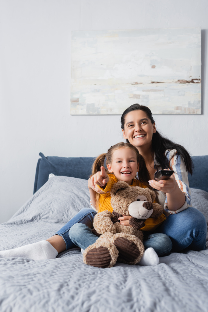excited girl pointing with finger while watching movie with mother and teddy bear - Photo, Image