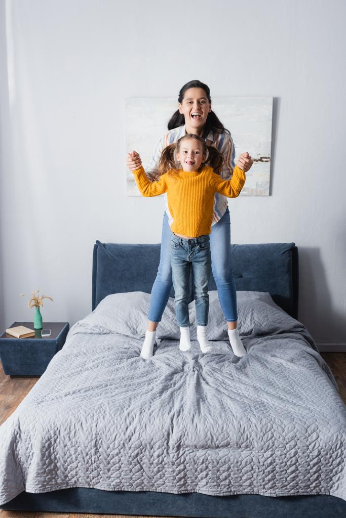 cheerful mother and daughter holding hands while jumping on bed - Photo, Image