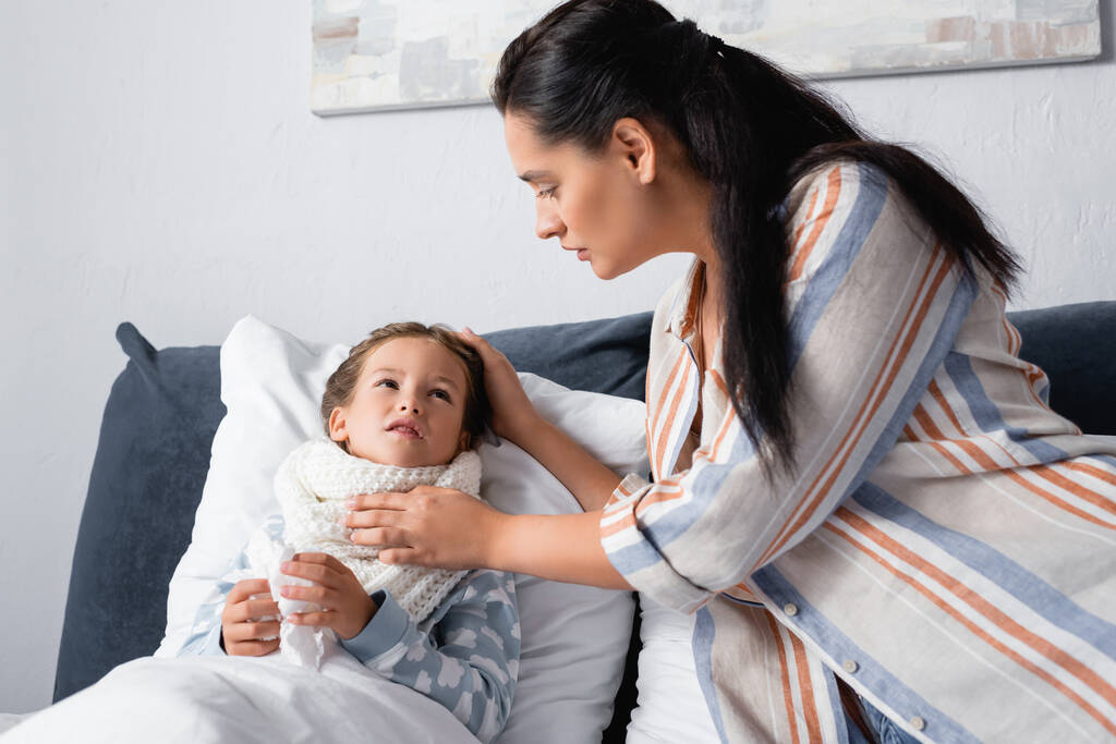 niño enfermo acostado en la cama, sosteniendo una servilleta de papel y mirando a la madre  - Foto, imagen