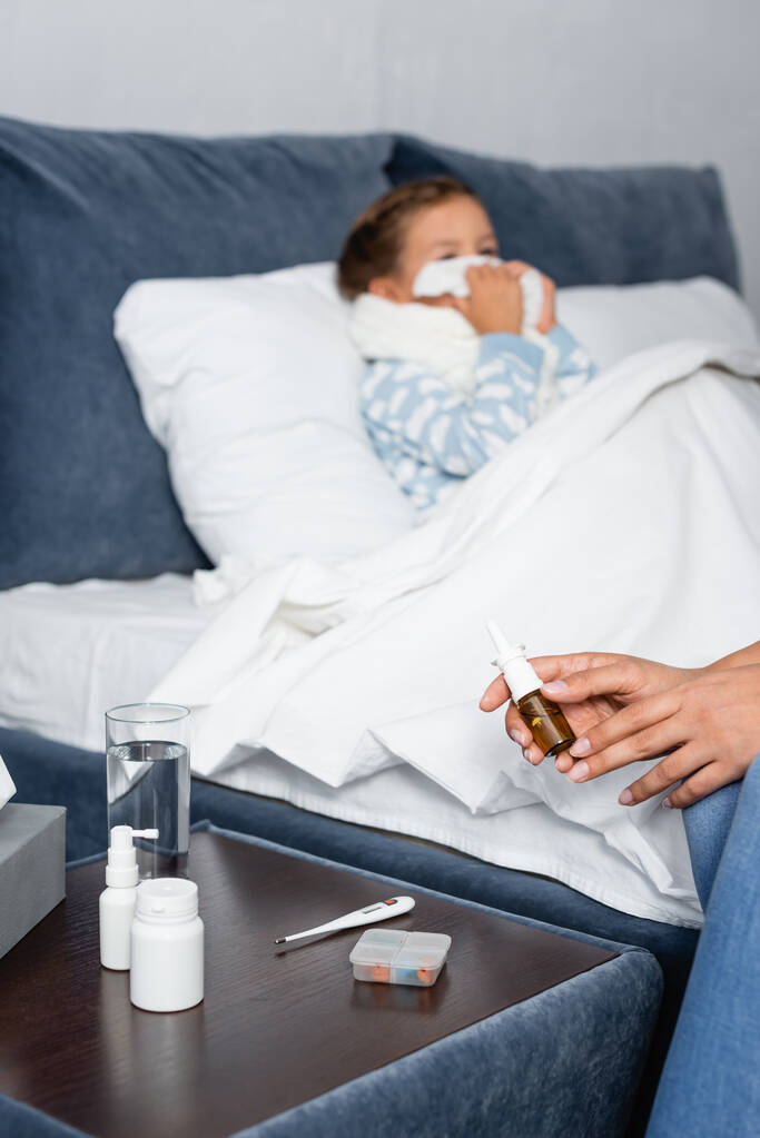 женщина с носовым спреем возле прикроватного столика с лекарствами и больной дочерью, лежащей в постели на размытом фоне - Фото, изображение