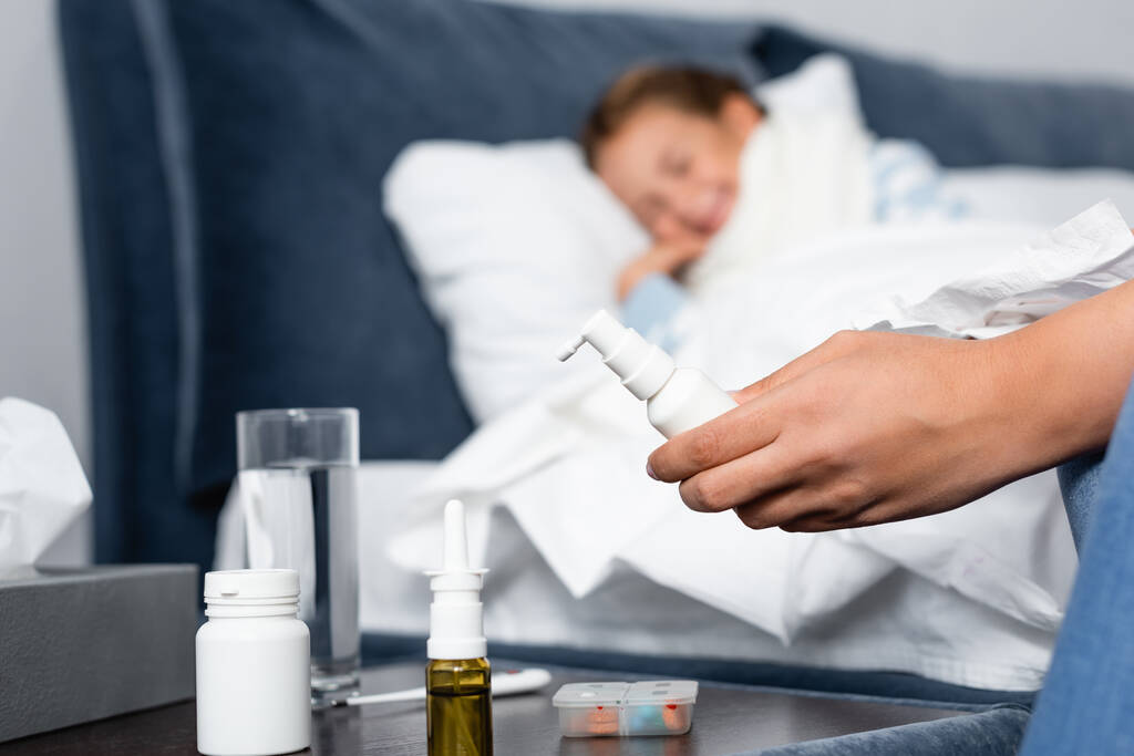 жінка тримає спрей для горла біля приліжкового столу з ліками та хворою дочкою, що лежить у ліжку на розмитому фоні
 - Фото, зображення