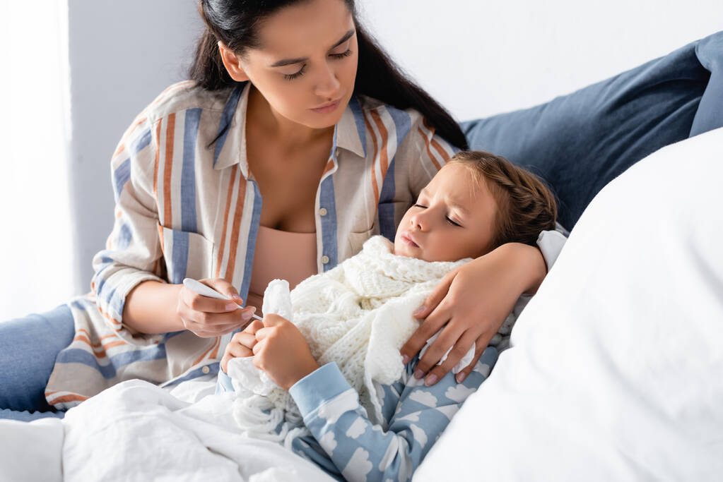 γυναίκα κρατώντας θερμόμετρο και αγκαλιάζοντας την άρρωστη κόρη ξαπλωμένη στο κρεβάτι με κλειστά μάτια - Φωτογραφία, εικόνα