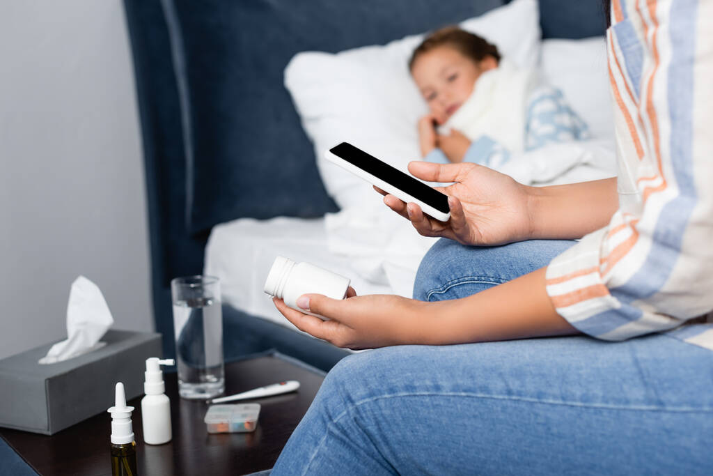 madre sosteniendo botella de pastillas y teléfono móvil cerca de hija enferma acostada en la cama - Foto, imagen
