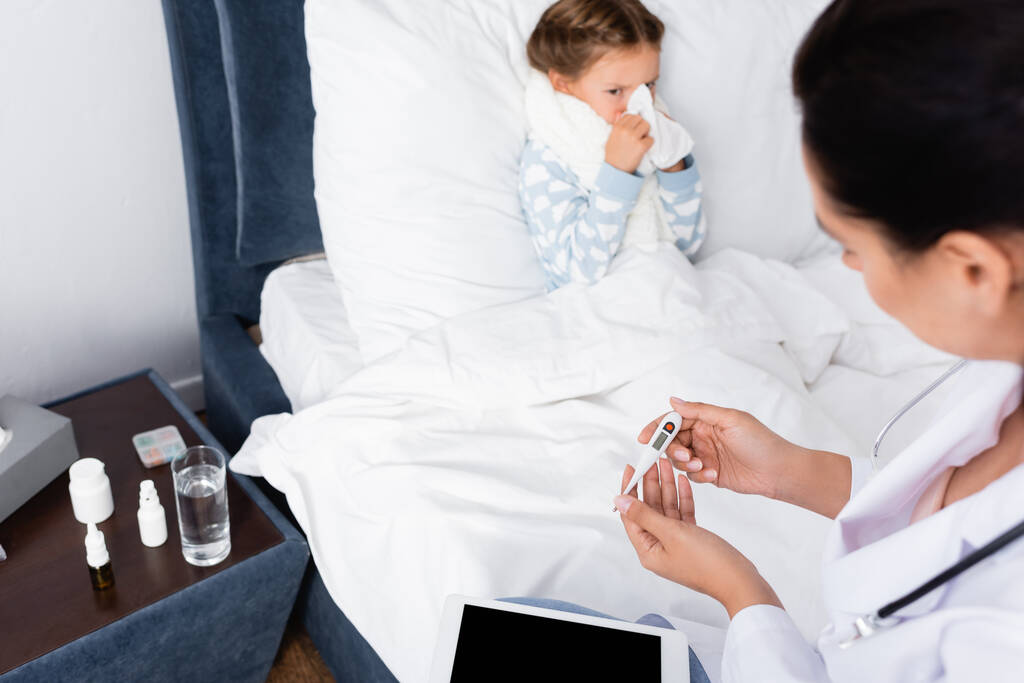 体温計を見ている医師の近くに紙ナプキンで鼻を拭く病気の女の子 - 写真・画像