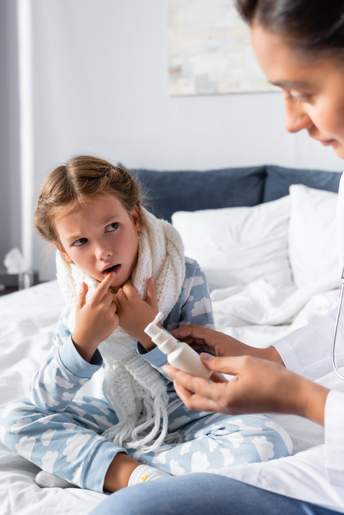 krankes Mädchen zeigt auf offenen Mund, während Arzt Halsspray hält - Foto, Bild