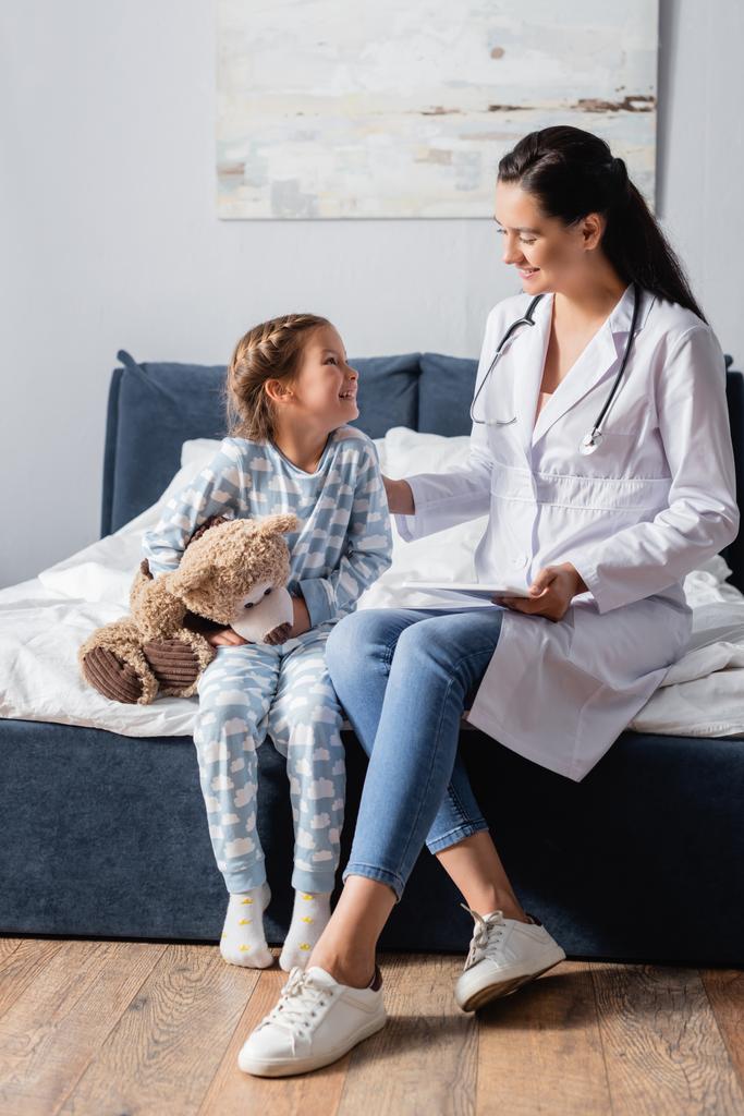 szczęśliwy dziecko z misiem patrząc na lekarza podczas siedzenia na łóżku - Zdjęcie, obraz