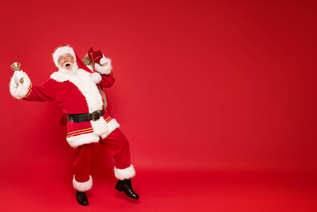 Счастливый, настоящий, забавный Санта Клаус держит мешок с рождественскими подарками, доставляет рождественские подарки. Красный фон, много места для копирования. - Фото, изображение