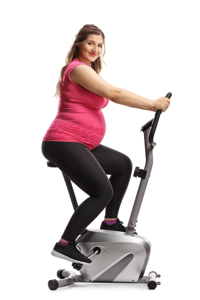 Pregnant woman in a pink top riding an exercise bike isolated on white background - Photo, Image